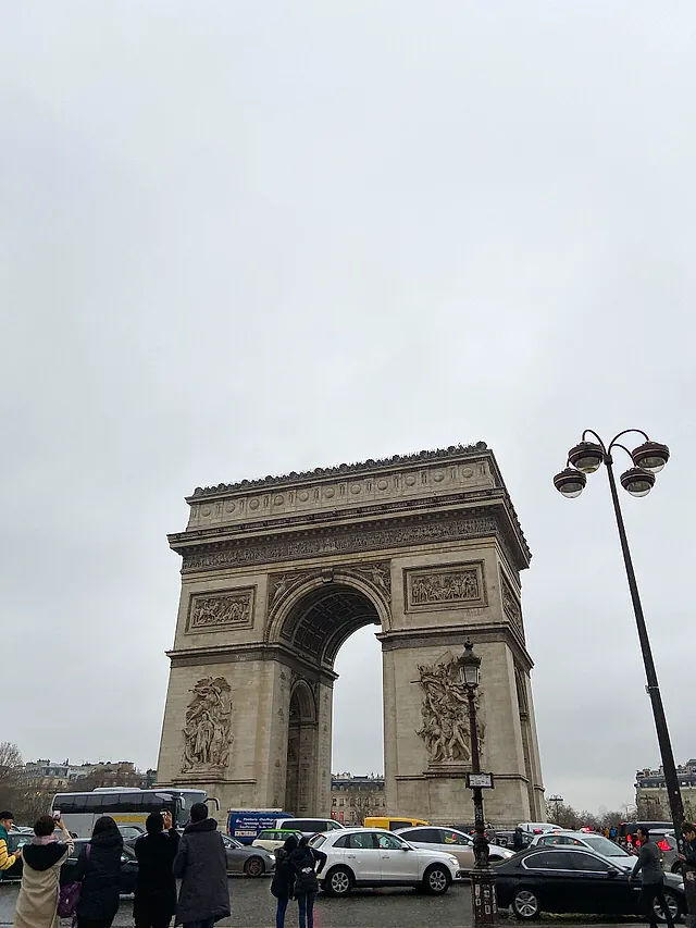 The Arc de Triomphe on a grey day, with a street lamp in the foreground and the sculpted attic above the arch.