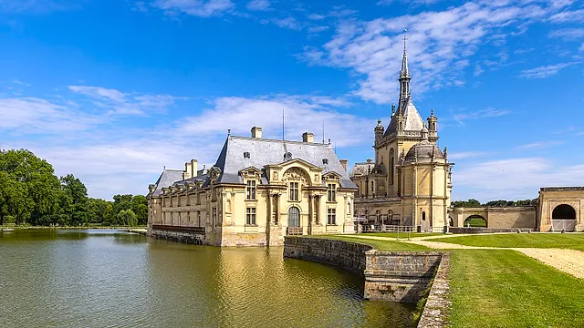 The Château de Chantilly standing at the edge of its water, with a slender tower and slate roofs under a blue sky with light clouds.