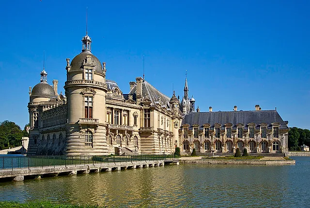 The Château de Chantilly rising from the water, with round towers, a domed turret and the long gallery wing, under a deep blue sky.