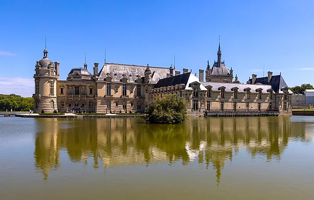The Château de Chantilly mirrored in the still water of its moat, seen from the paved road on a sunny day.