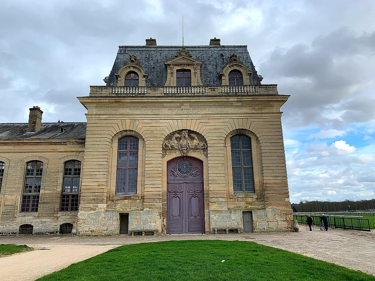The monumental stone front of the Grandes Écuries at Chantilly, with arched windows and a carved central pediment, above a green lawn.