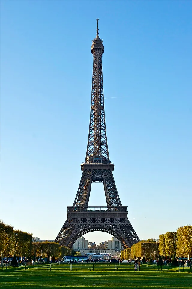 The Eiffel Tower seen from the Champ de Mars in a tall view, a slim iron silhouette against a clear blue sky.