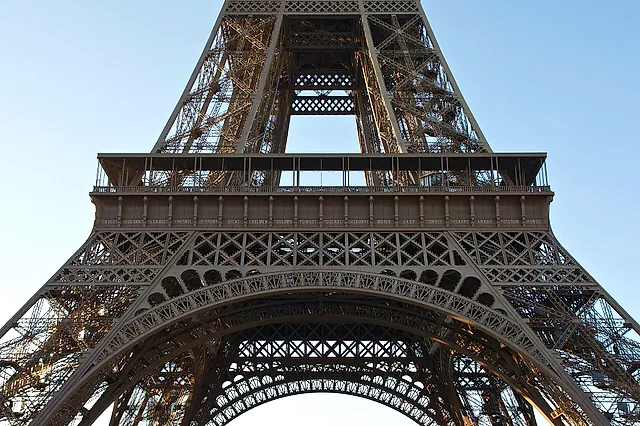 The lower part of the Eiffel Tower seen from the Champ de Mars, with the great arch and the first-floor platform against a pale blue sky.