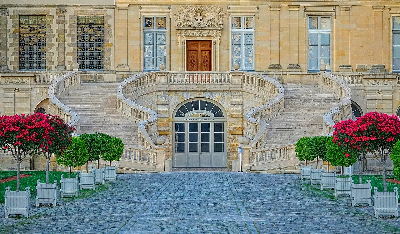 The curved double staircase at the Château de Fontainebleau, with flowering shrubs in white planters on either side and a cobbled courtyard in front.