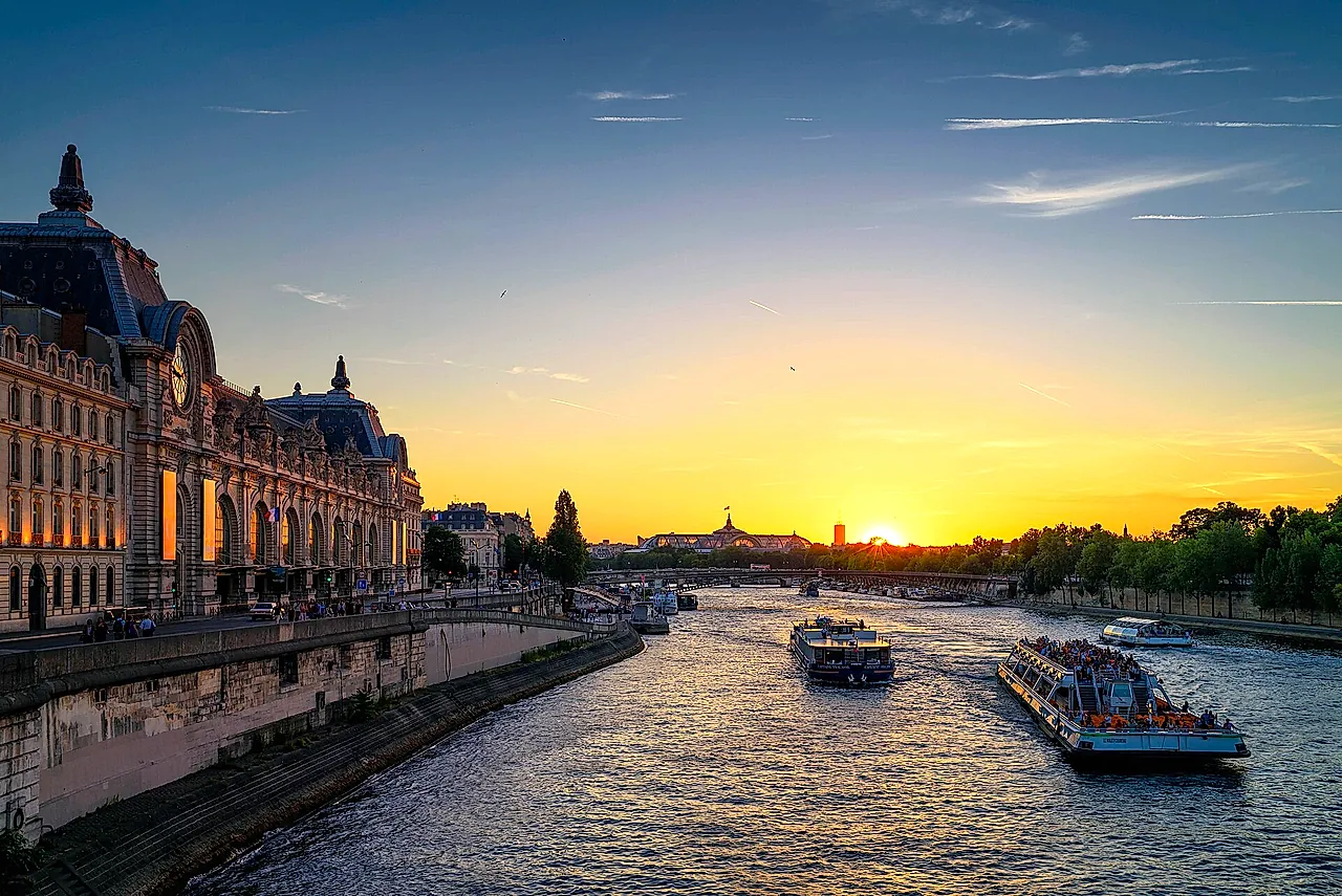 Sunset over the Seine beside the Musée d'Orsay, with the river glowing orange and two sightseeing boats passing under the bridges.
