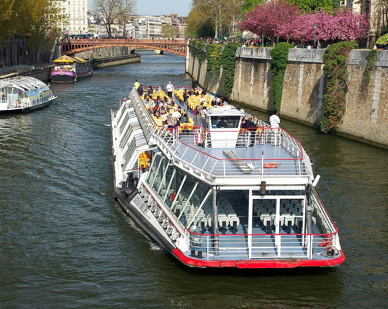 A glass-roofed sightseeing boat on the Seine near Notre-Dame, with passengers on the open upper deck and another boat behind.