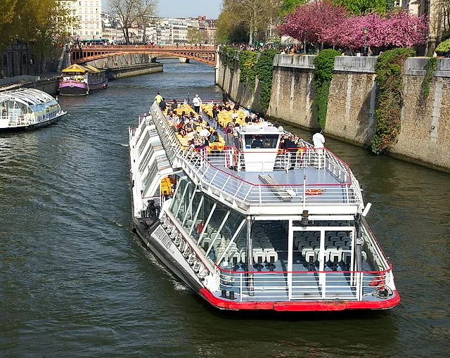 A glass-roofed sightseeing boat on the Seine near Notre-Dame, with passengers on the open upper deck and another boat behind.
