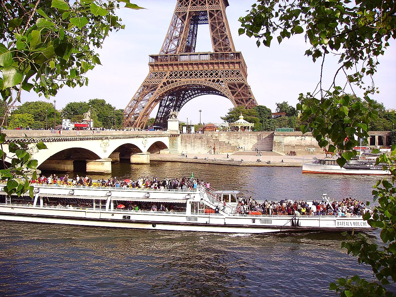 A sightseeing boat full of passengers on the Seine with a stone bridge and the top of the Eiffel Tower behind, seen through the trees.