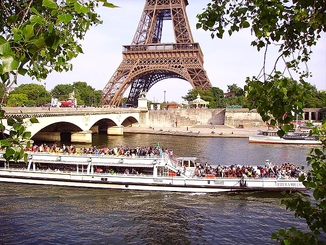 A sightseeing boat full of passengers on the Seine with a stone bridge and the top of the Eiffel Tower behind, seen through the trees.