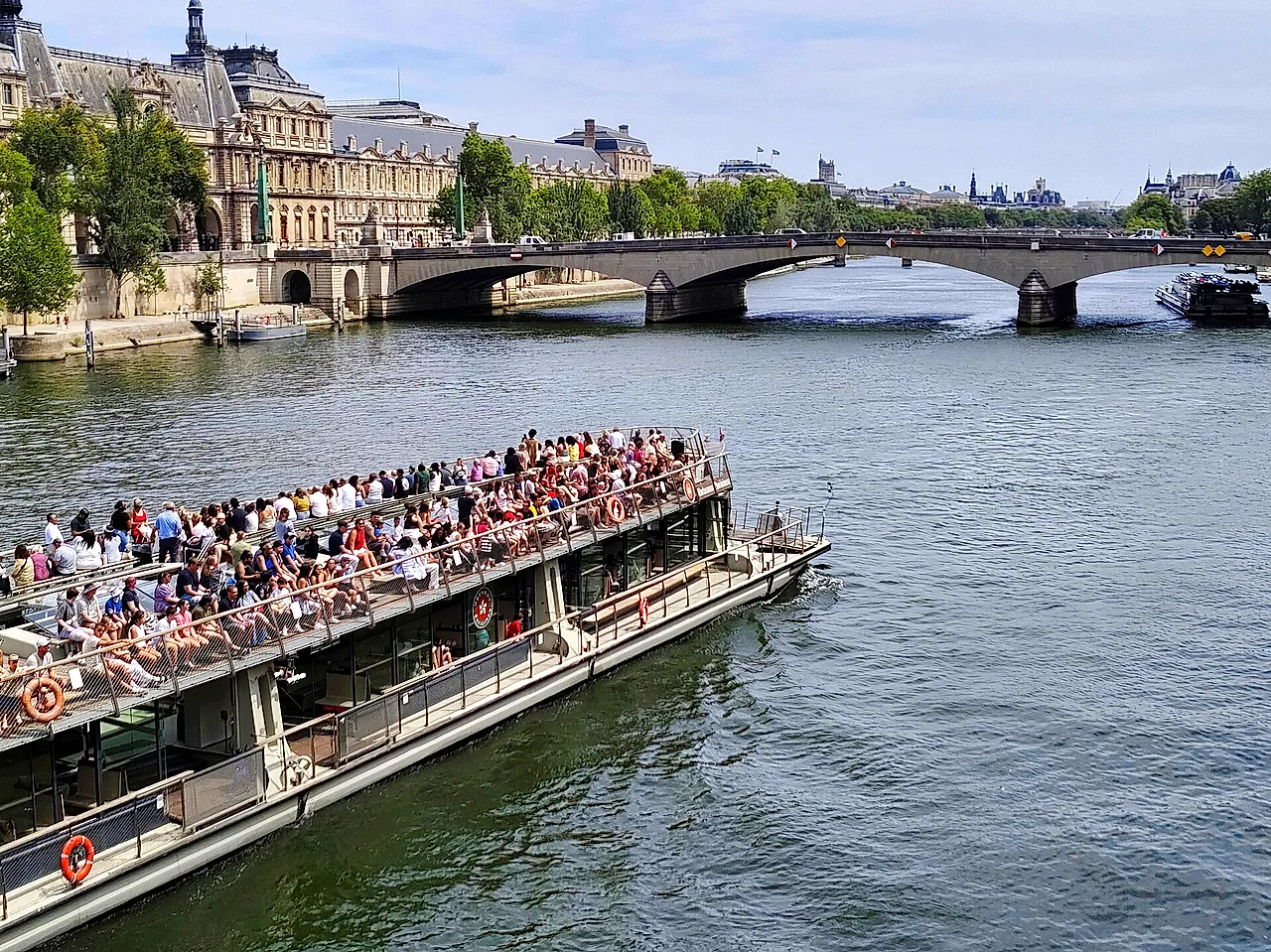 A crowded open-top tourist boat cruising on the Seine towards the Pont du Carrousel, with the old buildings of the left bank behind.