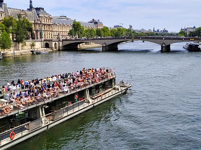 A crowded open-top tourist boat cruising on the Seine towards the Pont du Carrousel, with the old buildings of the left bank behind.