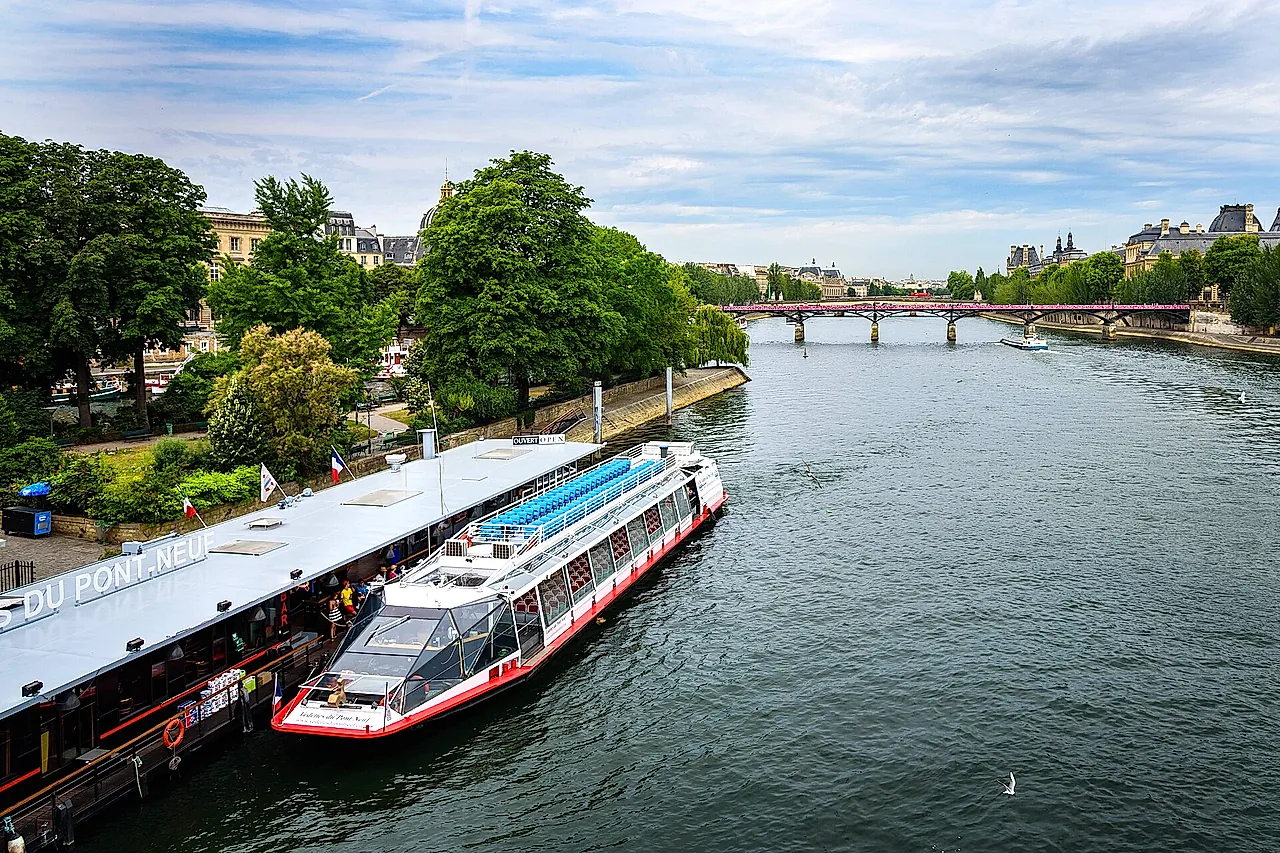 A white and blue sightseeing boat leaving its landing on the Seine, with trees on the bank and a bridge in the distance.