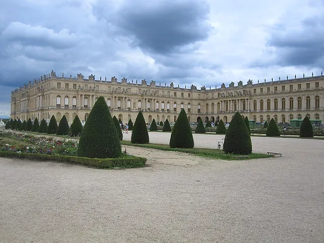 The long façade of the Palace of Versailles seen across the clipped hedges and gravel paths of the gardens, under a cloudy sky.